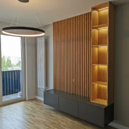 Nowoczesny salon z drewnianą ścianą z pionowych listew, szarą szafką RTV i podświetlanym regałem. Okrągła lampa sufitowa, parkiet i widok na balkon.