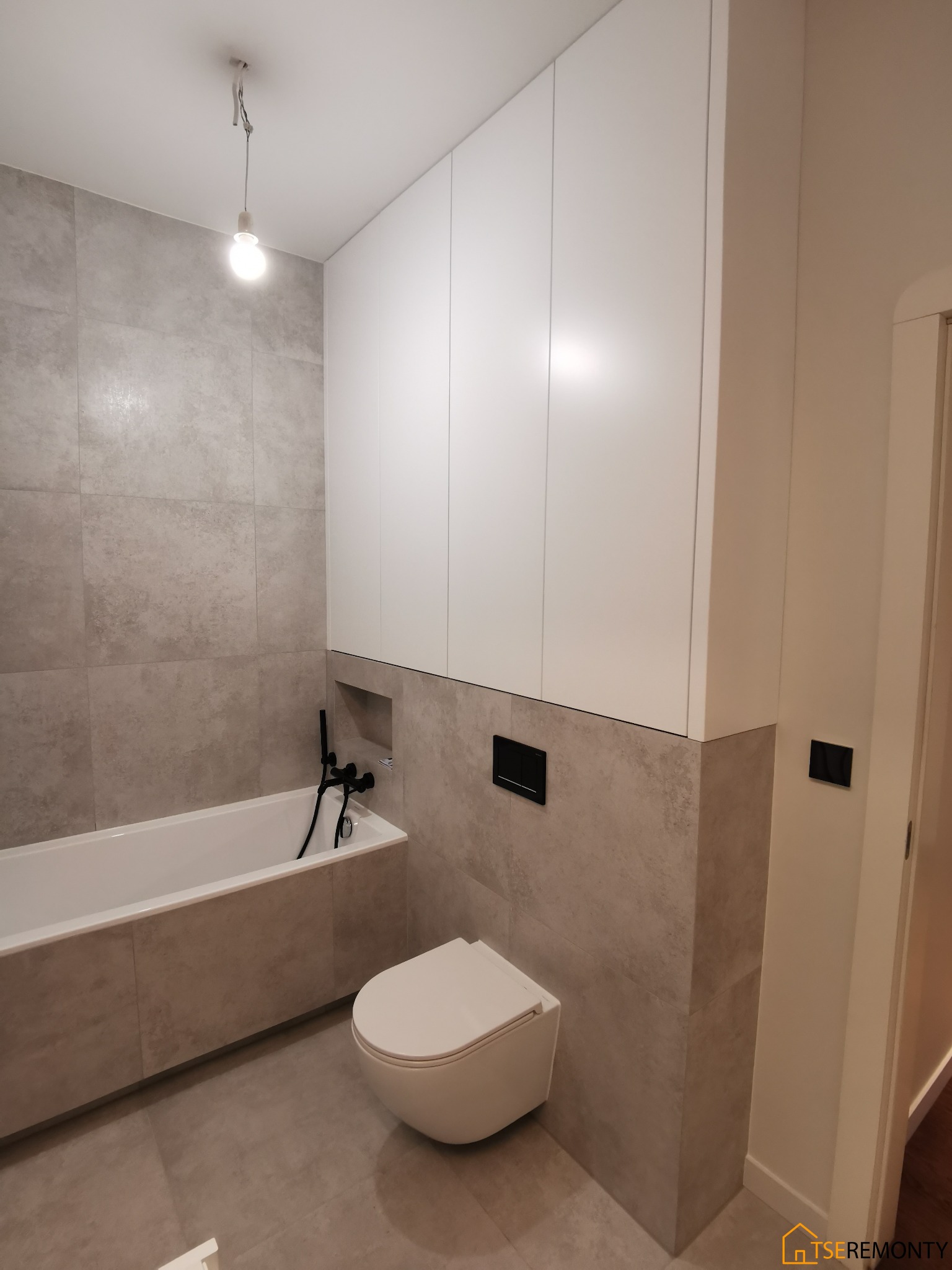 Nowoczesna łazienka w odcieniach szarości z białą wanną, toaletą wiszącą i szafkami zabudowanymi nad wanną, armatura w kolorze czarnym, surowa żarówka zwisająca z sufitu.