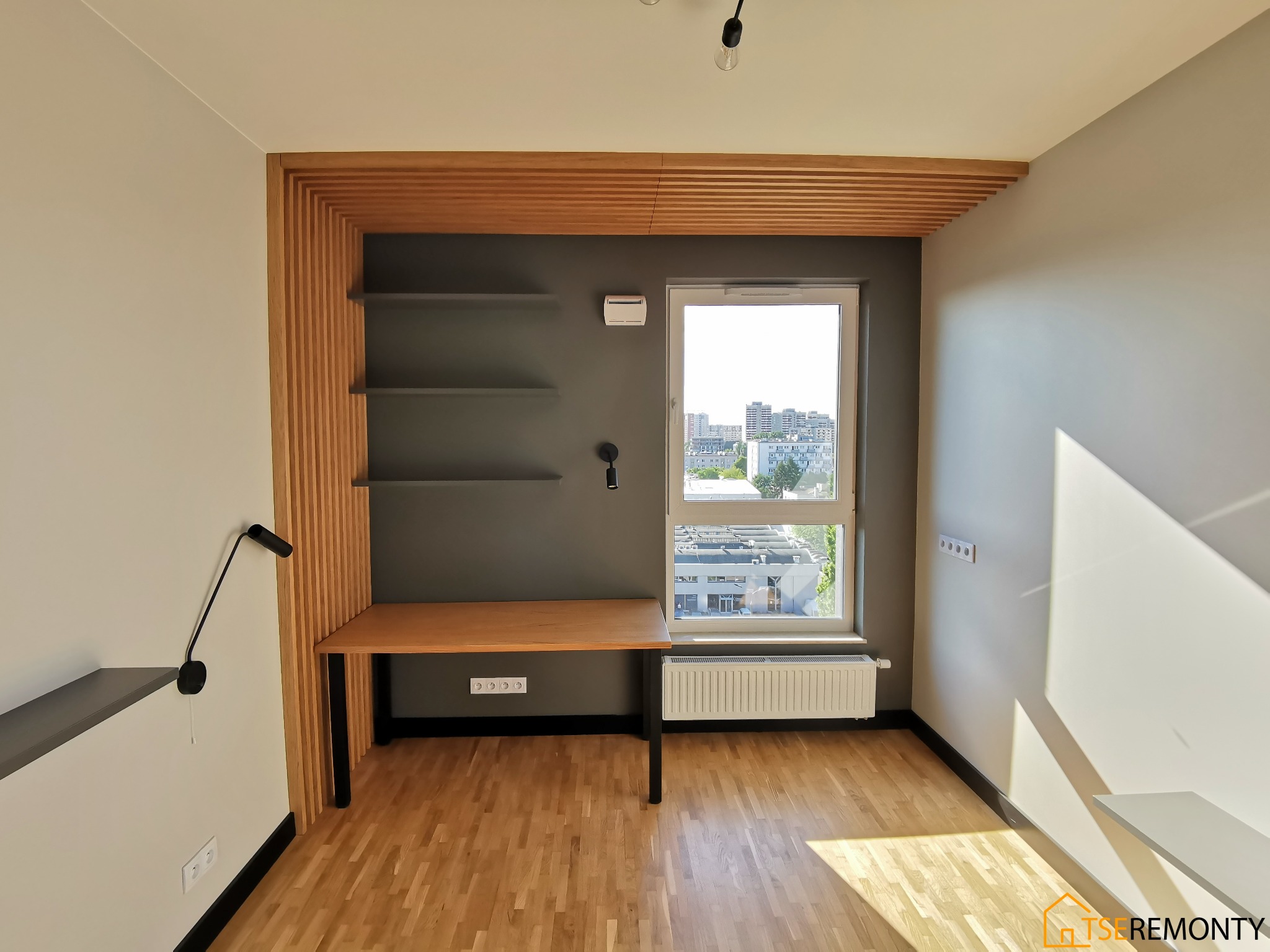 Pokój po remoncie: drewniana podłoga, szara ściana z półkami i biurkiem, drewniane lamele na ścianie i suficie, okno z widokiem na miasto.