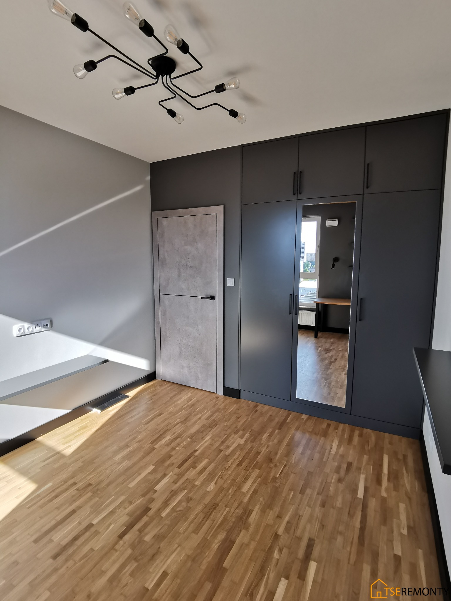 Szare wnętrze pokoju z drewnianą podłogą, zabudową szaf w kolorze grafitowym z lustrem, szarą ścianą i drzwiami oraz lampą sufitową w stylu pająka.