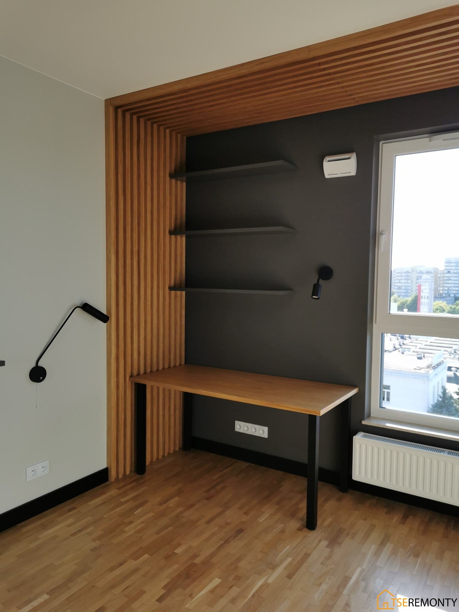 Drewniana zabudowa biurka z lamelami i półkami w pokoju z parkietem, widok na miasto przez okno.