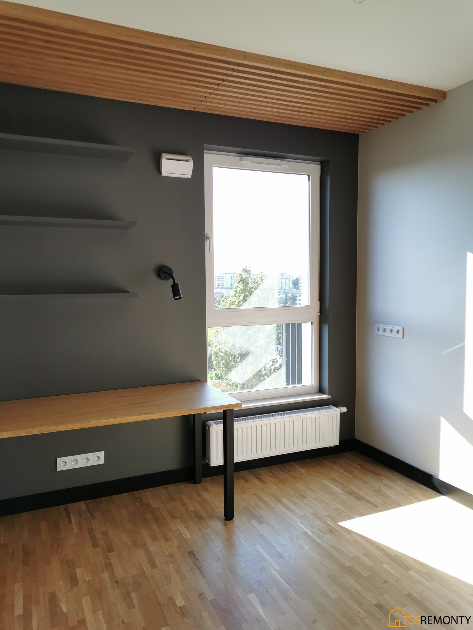 Pokój po remoncie: drewniana podłoga, szara ściana z półkami i biurkiem, drewniany sufit lamelowy, okno z grzejnikiem.