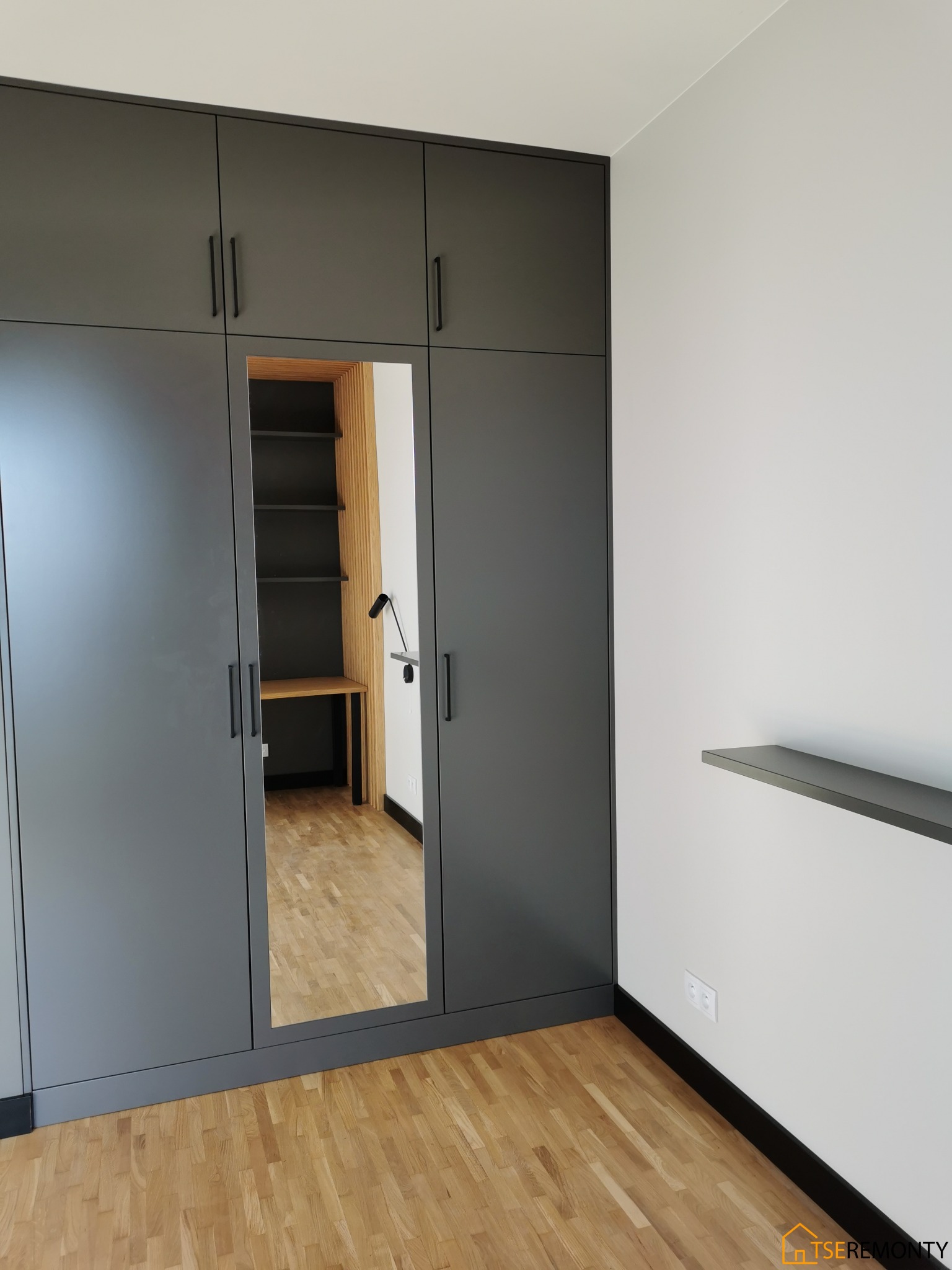 Szara szafa wnękowa z lustrzanym frontem, w której widać biurko z półkami i lampką, drewniana podłoga i listwy przypodłogowe.