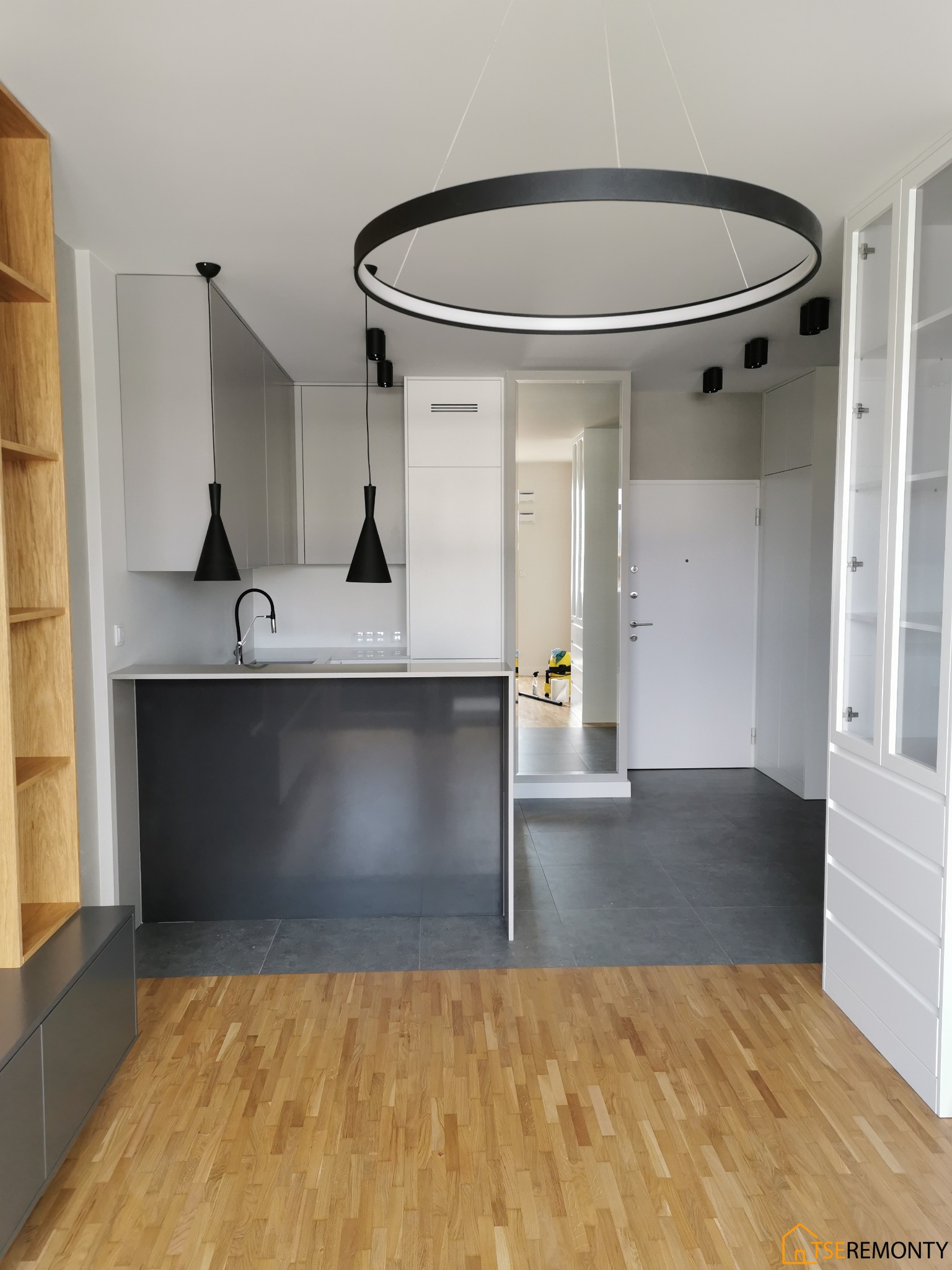 Nowoczesna kuchnia z wyspą, szare szafki, czarne lampy wiszące i okrągła lampa sufitowa, drewniana podłoga i szara podłoga w kuchni, widok na przedpokój z białymi drzwiami.