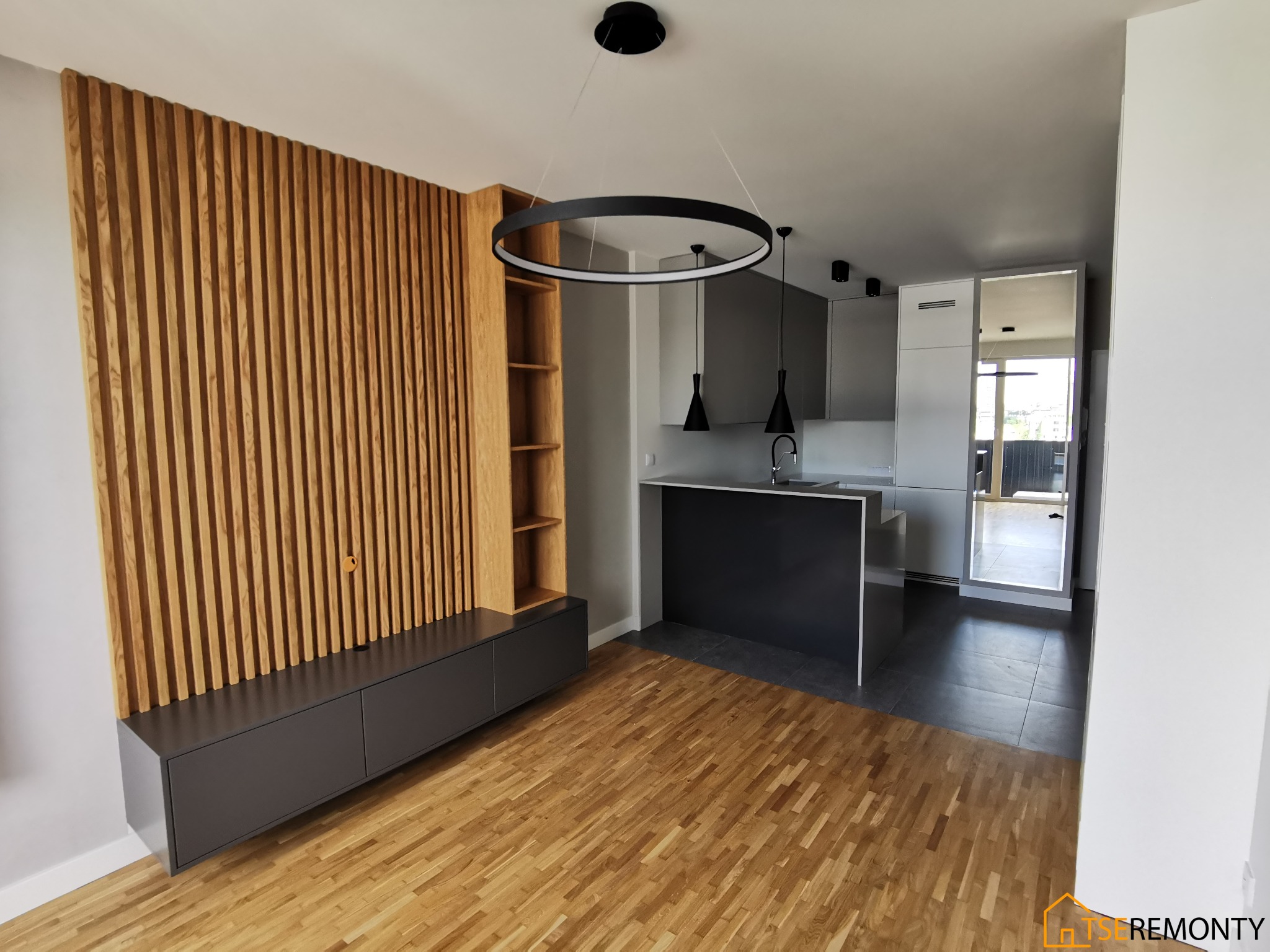 Nowoczesne wnętrze z drewnianą ścianką działową i szafką RTV, parkiet dębowy, otwarta kuchnia z szarymi szafkami i czarnym blatem, okrągła lampa wisząca.