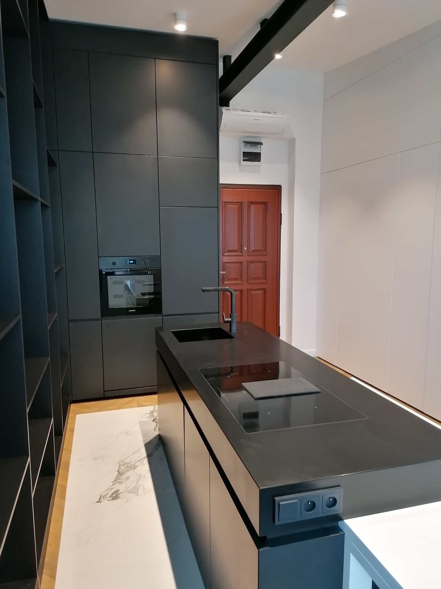 Nowoczesna kuchnia z czarnymi, matowymi frontami szafek, wyspą z płytą indukcyjną i marmurową podłogą, uzupełniona o drewniane drzwi w tle.