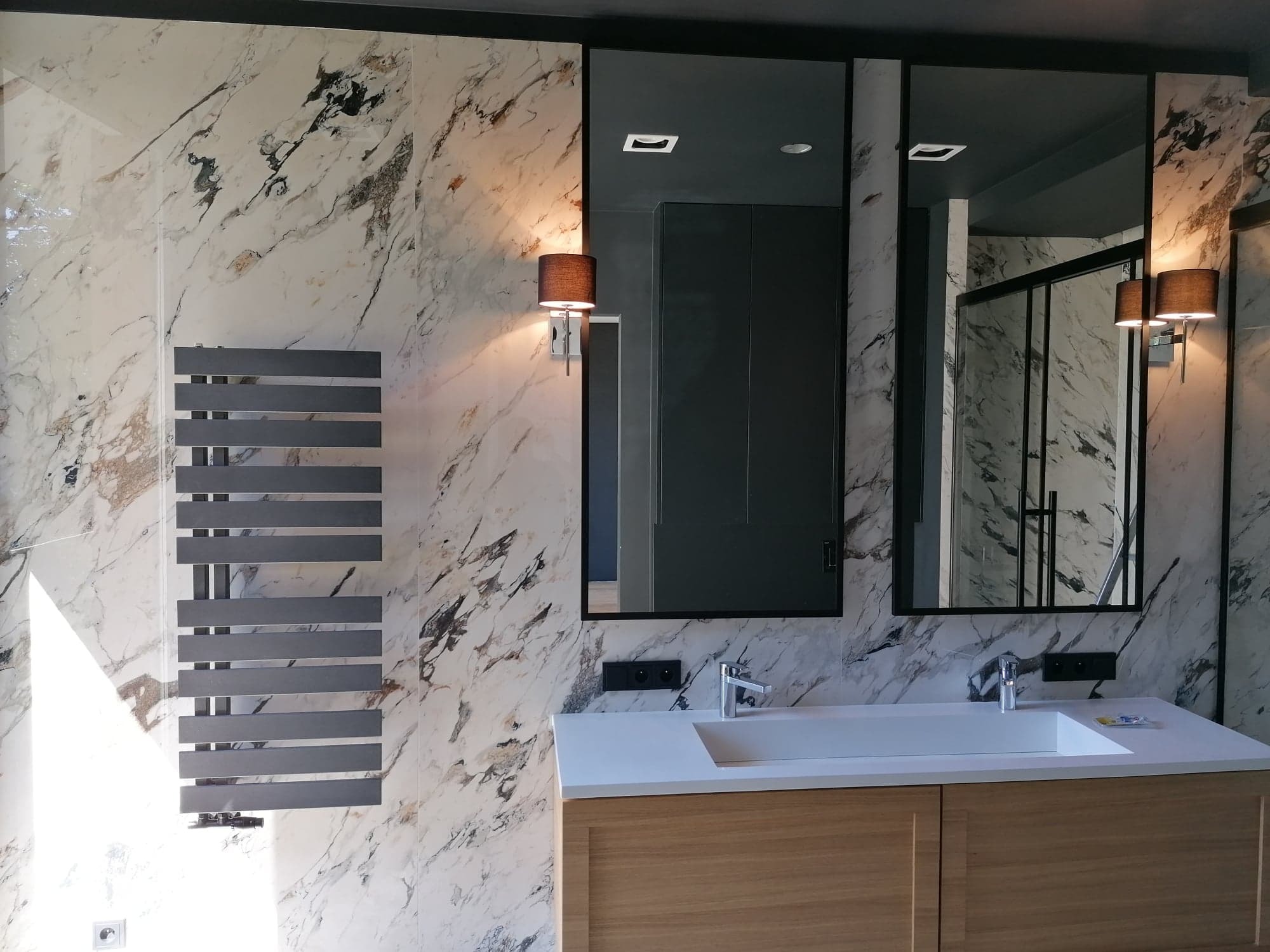 Nowoczesna łazienka z marmurową ścianą, grzejnikiem drabinkowym, dwiema lampami ściennymi z abażurami i szafką pod umywalkę z jasnego drewna.