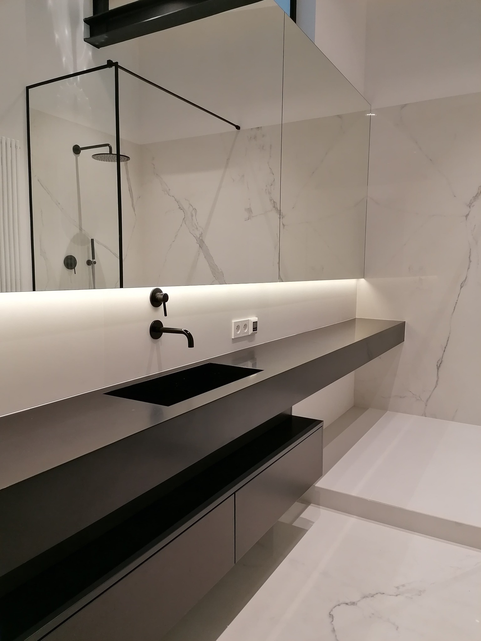 Nowoczesna łazienka z czarną armaturą, umywalką i szafką podumywalkową na tle białych marmurowych płytek i szklanej kabiny prysznicowej.