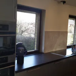 Szare szafki kuchenne z czarnym blatem, widok na okno z drzewami, piekarnik w zabudowie i lampa dekoracyjna na blacie.