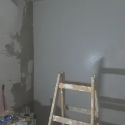 Fragment pokoju w trakcie remontu: ściana częściowo pokryta surowym tynkiem, częściowo pomalowana na szaro, drewniana drabina malarska, narzędzia i materiały na szafce.