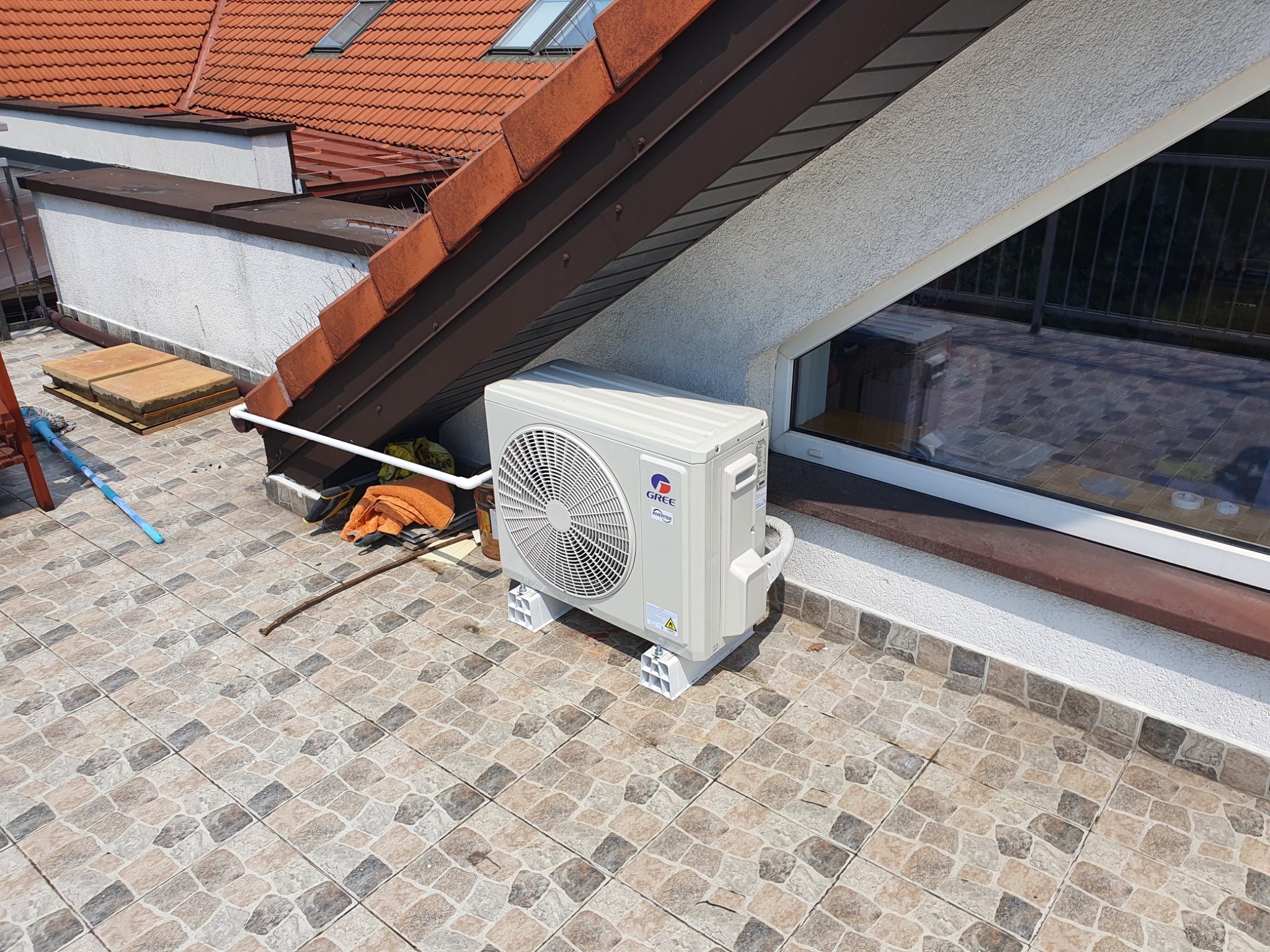 Zewnętrzna jednostka klimatyzacji marki Gree na dachu budynku z czerwoną dachówką, widoczna pod oknem.
