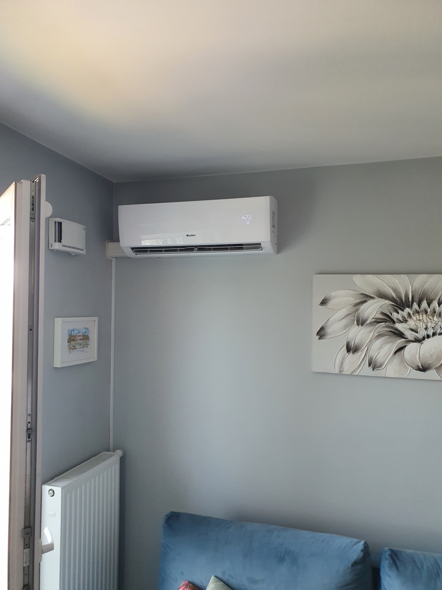 Biały klimatyzator ścienny z wyświetlaczem temperatury 23 stopnie, zamontowany w szarym pokoju obok obrazu z kwiatem, widoczny również grzejnik i fragment sofy.