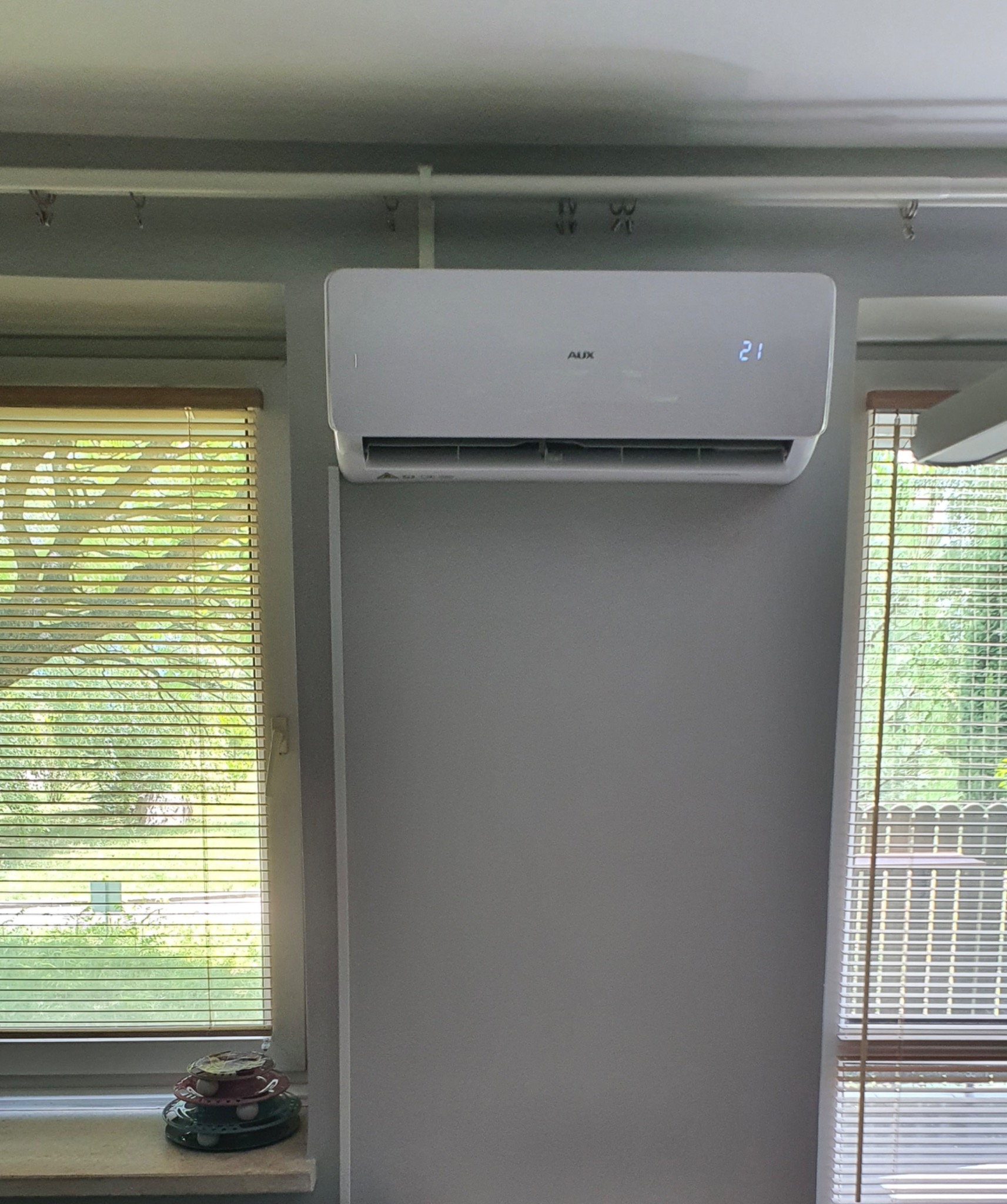 Biały klimatyzator marki AUX zamontowany na szarej ścianie, obok okna z żaluzjami, wyświetlający temperaturę 21 stopni.