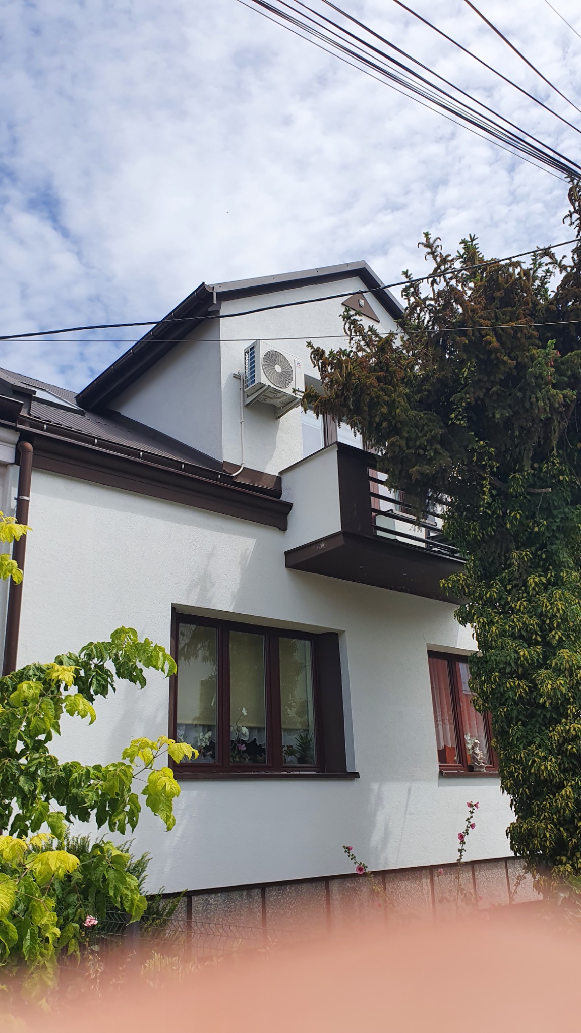 Biały budynek z brązowymi oknami i balkonem, na ścianie zamontowana jednostka zewnętrzna klimatyzatora, widoczne drzewa i niebo.
