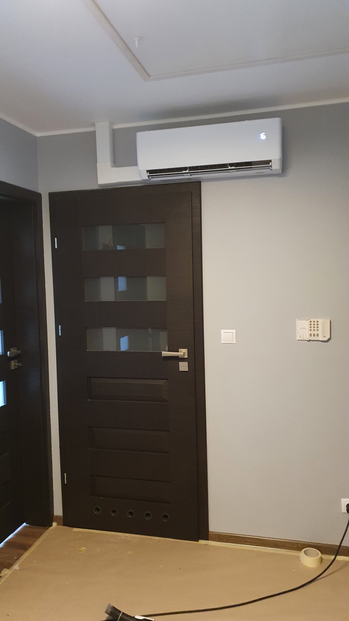 Biały klimatyzator ścienny zamontowany nad ciemnymi drzwiami z przeszkleniami, widoczna instalacja elektryczna i przewody na podłodze.