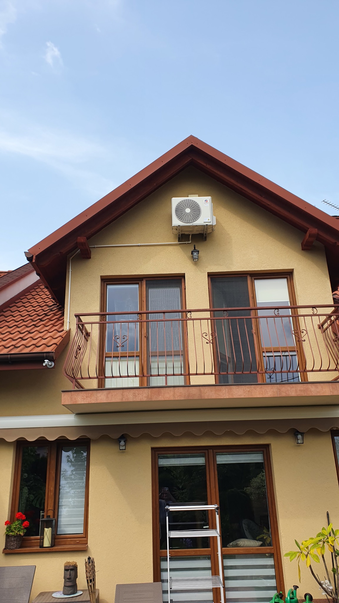 Biały klimatyzator zamontowany na żółtej elewacji domu z brązowym dachem i balkonem z ozdobną balustradą, widoczna drabina oparta o ścianę.