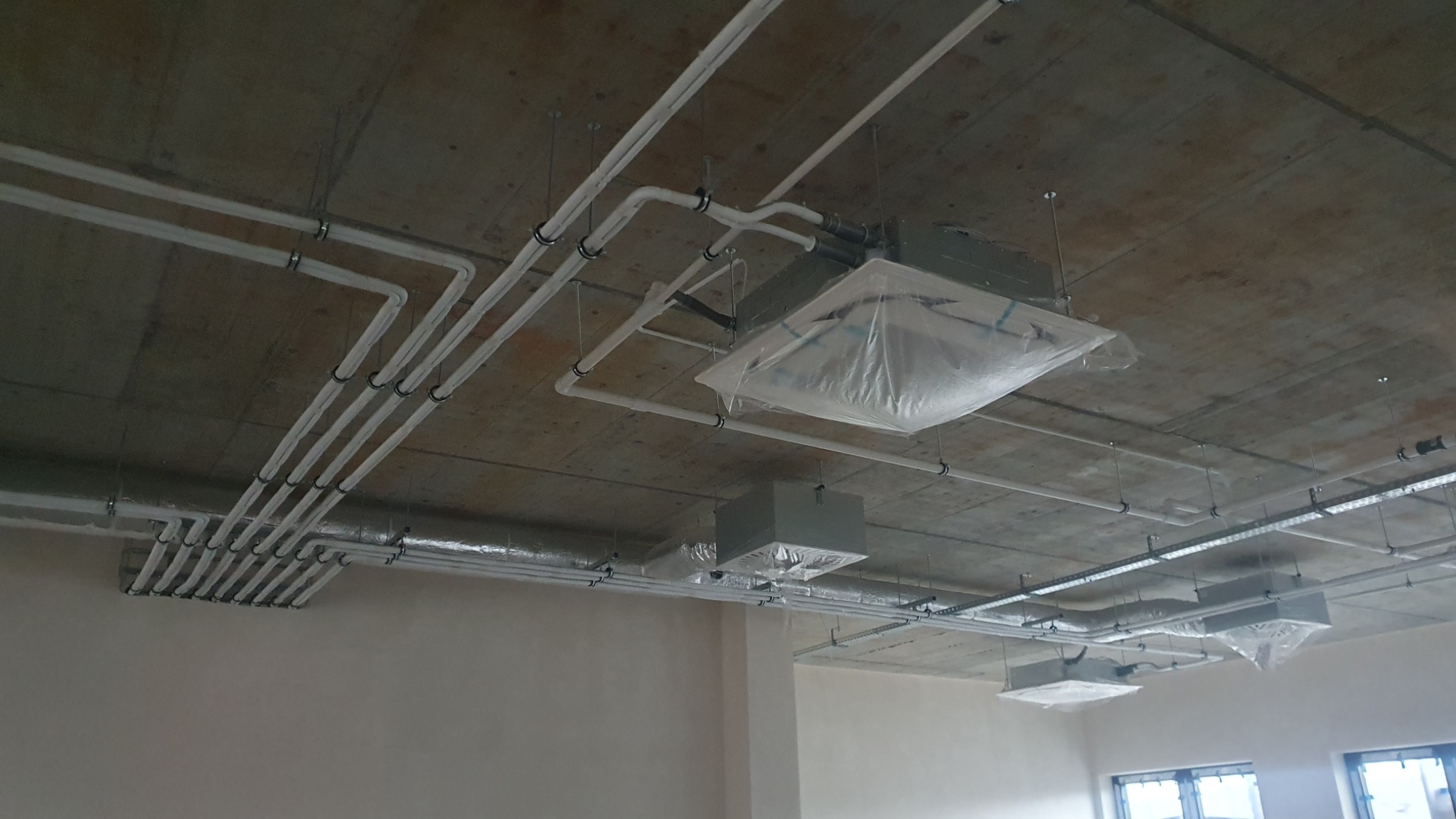 Instalacja przewodów wentylacyjnych i jednostek klimatyzacyjnych pod surowym betonowym sufitem, zabezpieczone folią ochronną.
