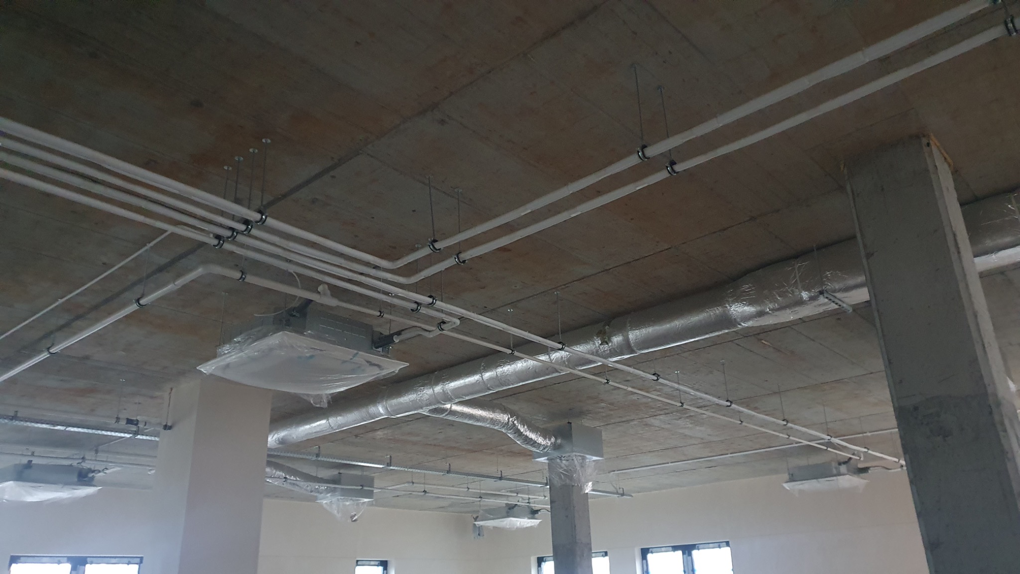 Instalacja wentylacyjna i rury pod sufitem z betonu, elementy zabezpieczone folią, widoczne okna w tle.