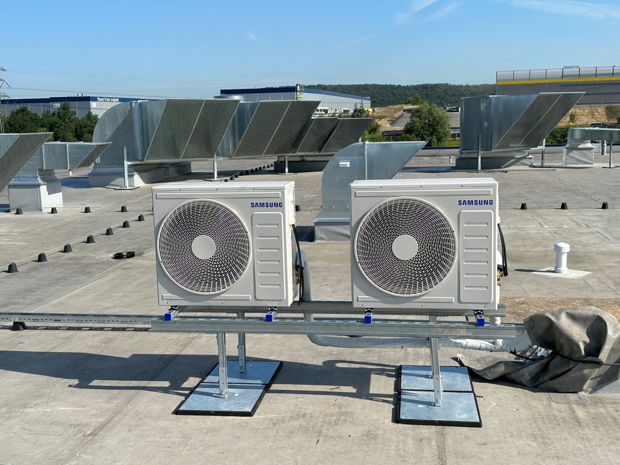 Dwa białe, zewnętrzne moduły Samsung na płaskim dachu, zamontowane na metalowej konstrukcji, z widocznymi przewodami i elementami wentylacji w tle.