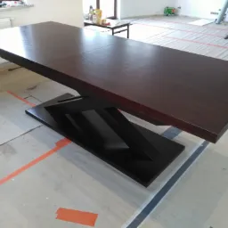 Rozłożony stół z ciemnego drewna na oryginalnej, czarnej, geometrycznej podstawie, w trakcie prac wykończeniowych w pomieszczeniu, podłoga zabezpieczona folią i taśmą.