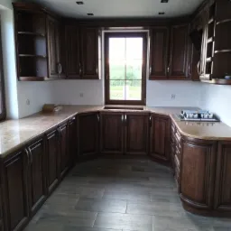 Klasyczna kuchnia z ciemnobrązowymi drewnianymi szafkami, marmurowym blatem i podłogą z płytek imitujących drewno, oświetlona wpuszczanymi lampami halogenowymi.