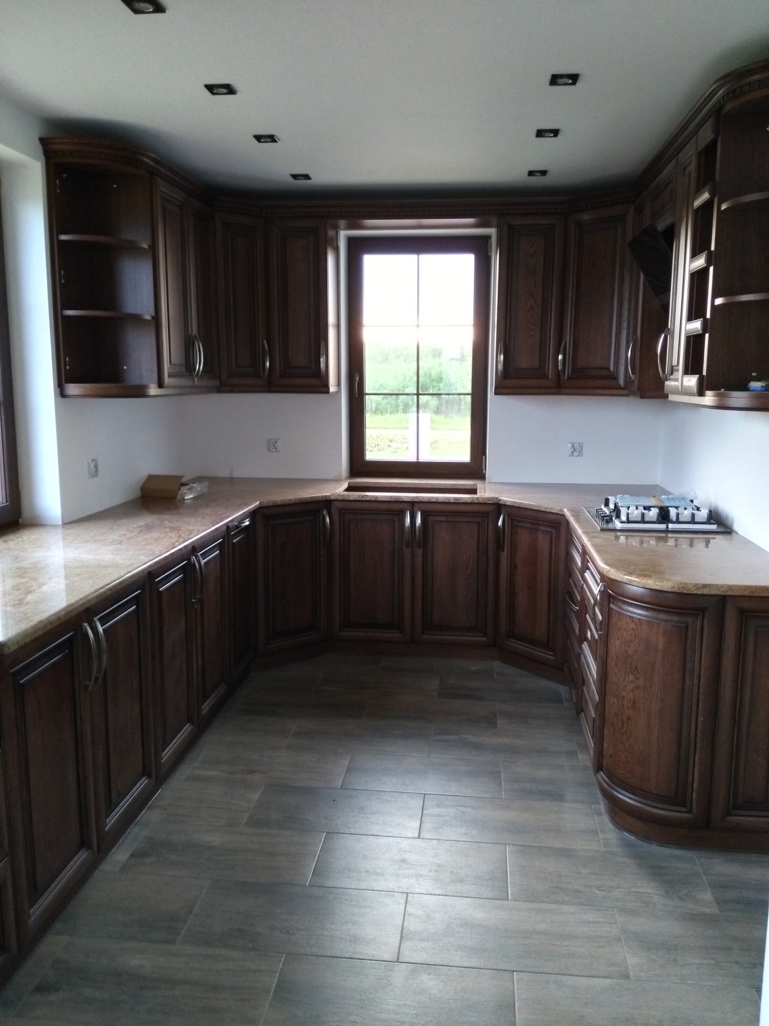 Klasyczna kuchnia z ciemnobrązowymi drewnianymi szafkami, marmurowym blatem i podłogą z płytek imitujących drewno, oświetlona wpuszczanymi lampami halogenowymi.