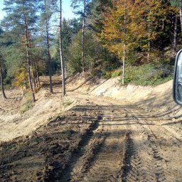 TRANS-KOP Tomasz Chudy - Ukształtowany teren leśny z widocznymi śladami opon ciężkiego sprzętu. W tle wysokie drzewa iglaste i liściaste w jesiennej kolorystyce. Lekko pofałdowany teren.