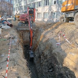 TRANS-KOP Tomasz Chudy - Pomarańczowa koparka w trakcie prac ziemnych, głęboki wykop z zabezpieczeniami, w tle budynek i inne maszyny budowlane. Prace ziemne w toku.