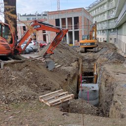 TRANS-KOP Tomasz Chudy - Pomarańczowe koparki JCB przy głębokim wykopie zabezpieczonym szalunkiem, w tle budynek w trakcie budowy i żuraw. Widoczny właz kanalizacyjny.