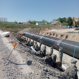 TRANS-KOP Tomasz Chudy - Długa, czarna rura karbowana w wykopie, zabezpieczona betonowymi podporami. W tle zabudowa jednorodzinna i koparka. Widoczne elementy geowłókniny.