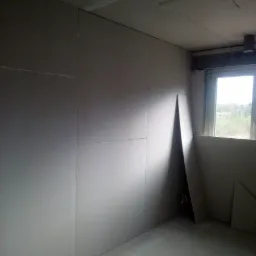 Wnętrze w trakcie montażu płyt kartonowo-gipsowych na ścianach i suficie, widoczne łączenia płyt i wystające przewody elektryczne, z oknem w tle.