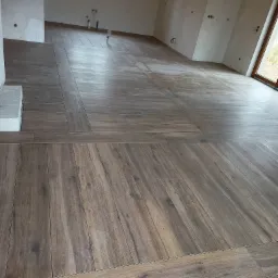 Ułożona podłoga z drewnopodobnych płytek ceramicznych w dużym, surowym pomieszczeniu z widocznymi instalacjami elektrycznymi i hydraulicznymi na ścianach.