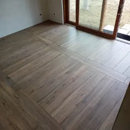 Ułożona podłoga z paneli imitujących drewno w jodełkę, widoczne ściany w stanie surowym i przesuwne drzwi balkonowe z widokiem na zewnątrz.