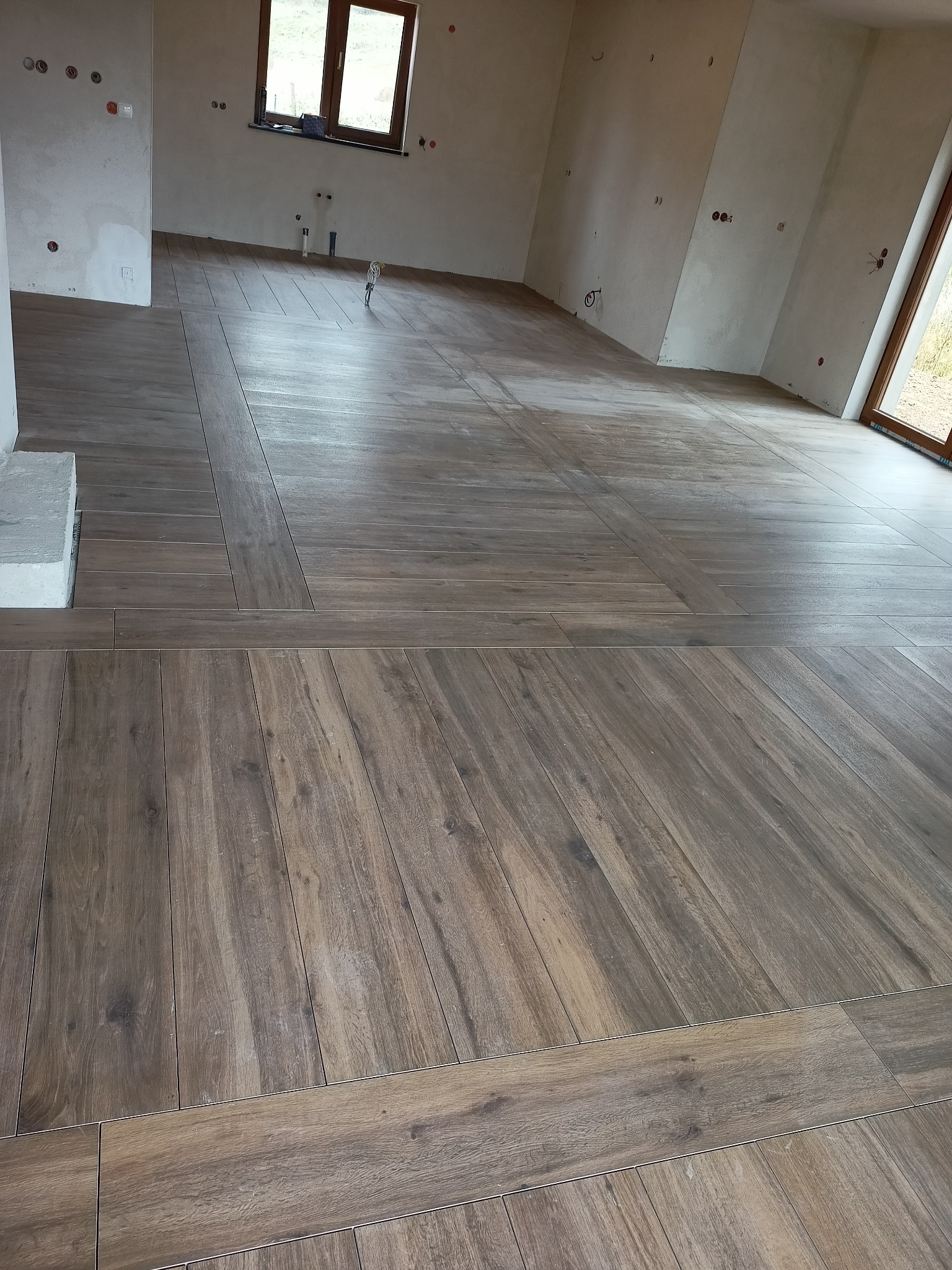 Ułożona podłoga z drewnopodobnych płytek ceramicznych w dużym, surowym pomieszczeniu z widocznymi instalacjami elektrycznymi i hydraulicznymi na ścianach.