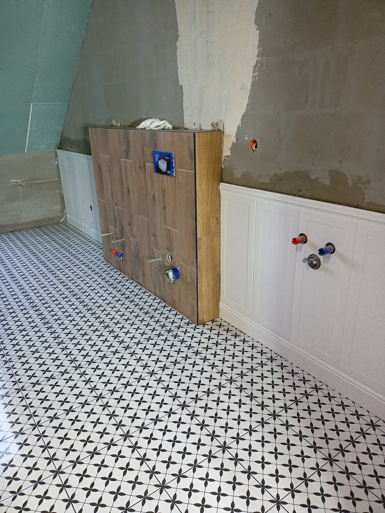 Wnętrze łazienki w trakcie remontu: zabudowa stelaża WC z płytkami imitującymi drewno, biała boazeria angielska, podłoga w czarno-białą geometryczną mozaikę, ściany częściowo w stanie surowym...