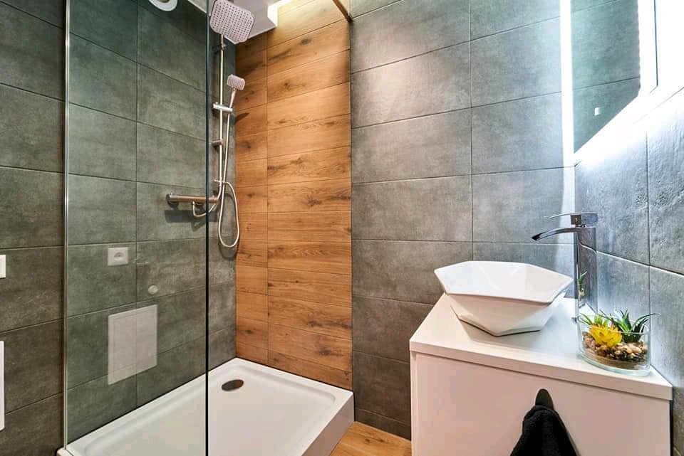 Nowoczesna łazienka z kabiną prysznicową ze szklaną ścianką, drewnopodobnymi panelami na ścianie i umywalką nablatową o geometrycznym kształcie, podświetlane lustro, szare płytki na ścianach.