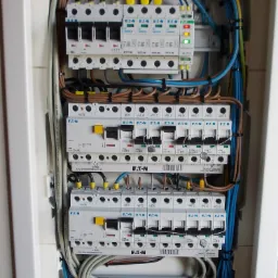 Otwarta skrzynka rozdzielcza z zainstalowanymi bezpiecznikami EATON, przewodami w kolorach niebieskim, brązowym i żółto-zielonym, oraz listwą zasilającą z diodami LED, zamontowana na ścianie z płytek.