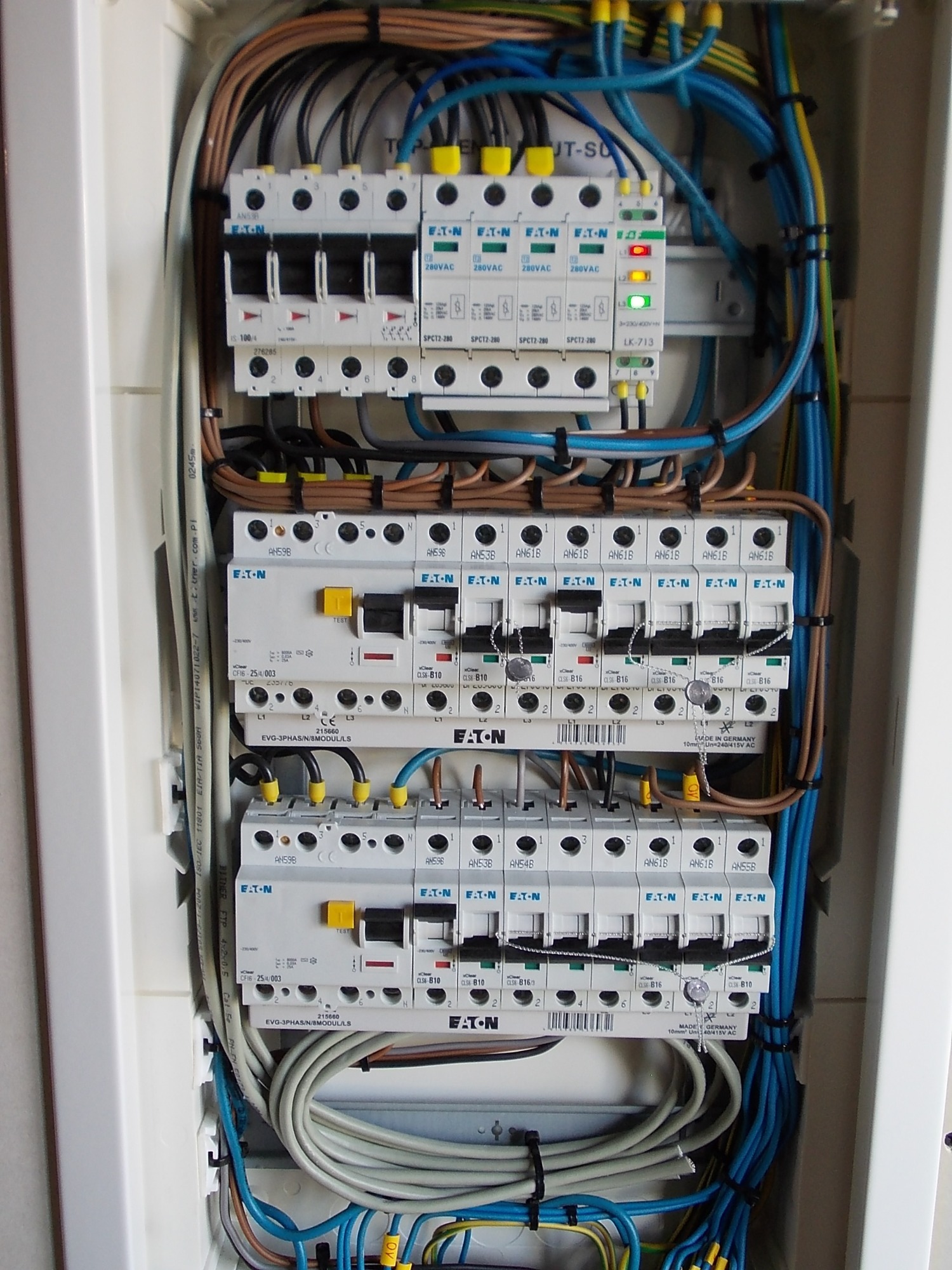 Otwarta skrzynka rozdzielcza z zainstalowanymi bezpiecznikami EATON, przewodami w kolorach niebieskim, brązowym i żółto-zielonym, oraz listwą zasilającą z diodami LED, zamontowana na ścianie z płytek.
