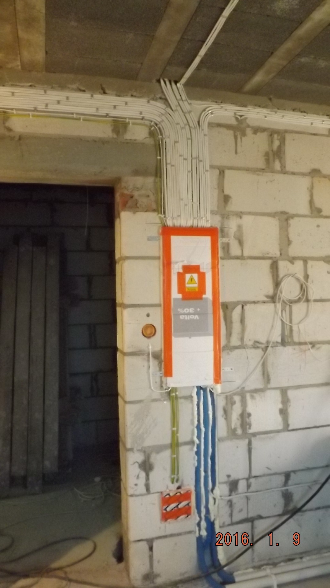 Rozdzielnica elektryczna Volta +30% z licznymi przewodami w białych rurkach zamocowanymi do sufitu i ściany z bloczków, widoczne gniazdko i otwór drzwiowy w tle.