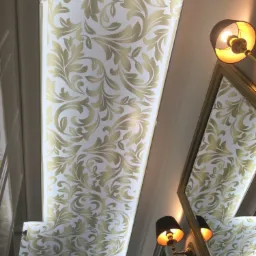 Biały sufit z dekoracyjnym złotym wzorem roślinnym, oświetlony lampami ściennymi, odbijający się w lustrze w złotej ramie.