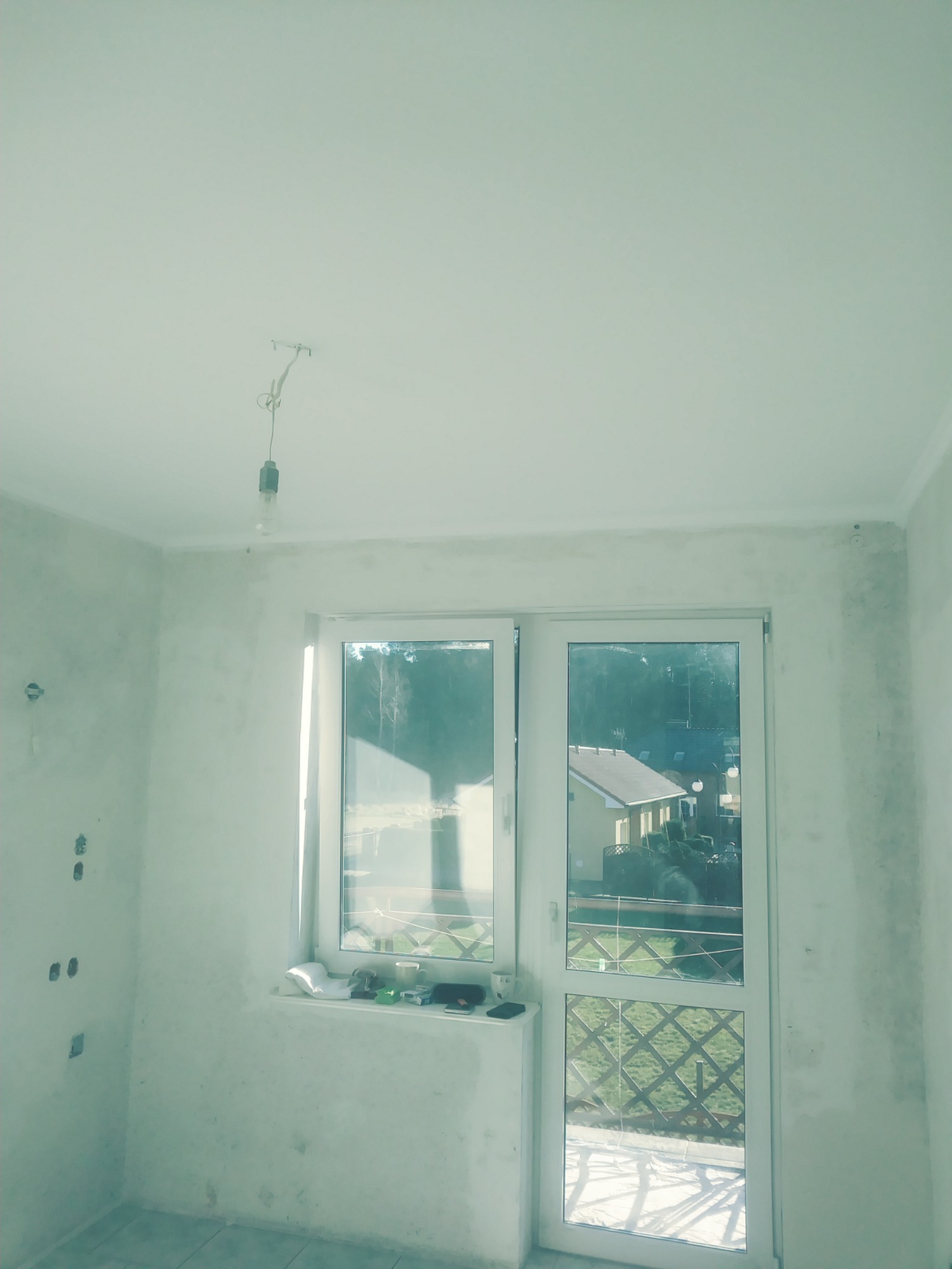 Pokój w trakcie przygotowania do prac wykończeniowych: ściany wstępnie zagruntowane, widoczne puszki elektryczne, okno balkonowe z widokiem na ogród, żarówka na kablu zwisająca z sufitu.