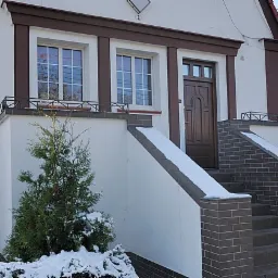 Wejście do domu z brązową, drewnianą konstrukcją, schodami z cegły i małym drzewkiem iglastym w śniegu.