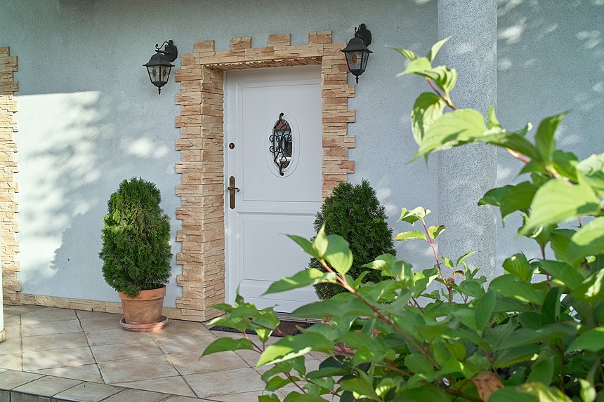 Białe wejściowe skrzydło z owalnym przeszkleniem, otoczone dekoracyjną okładziną z imitacji kamienia, oświetlone kinkietami w stylu retro, z roślinami w donicach po bokach.