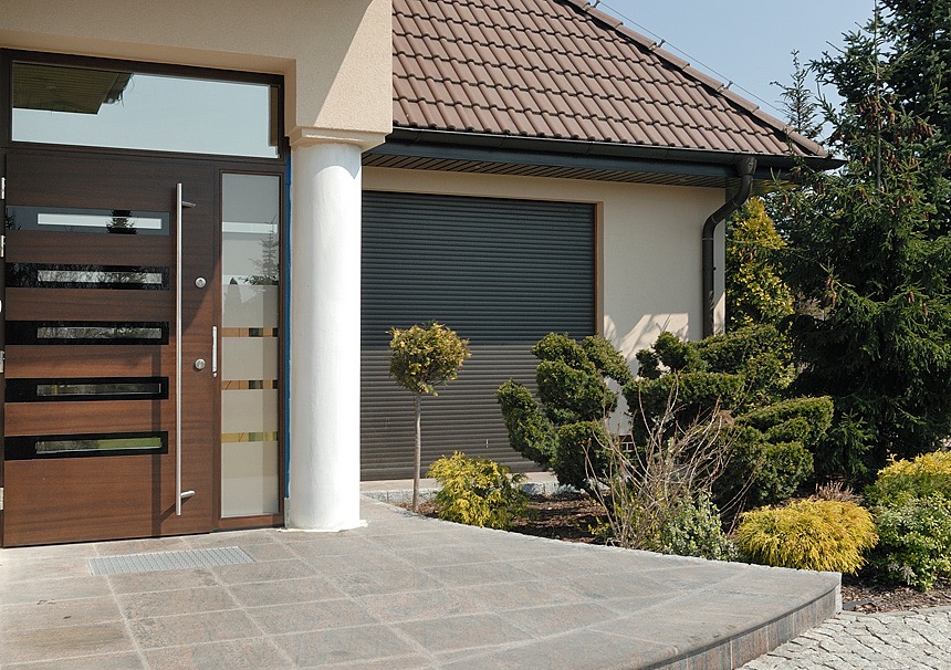 Nowoczesne wejście do domu z brązowymi skrzydłami z przeszkleniami, białą kolumną i szarą bramą garażową roletową.