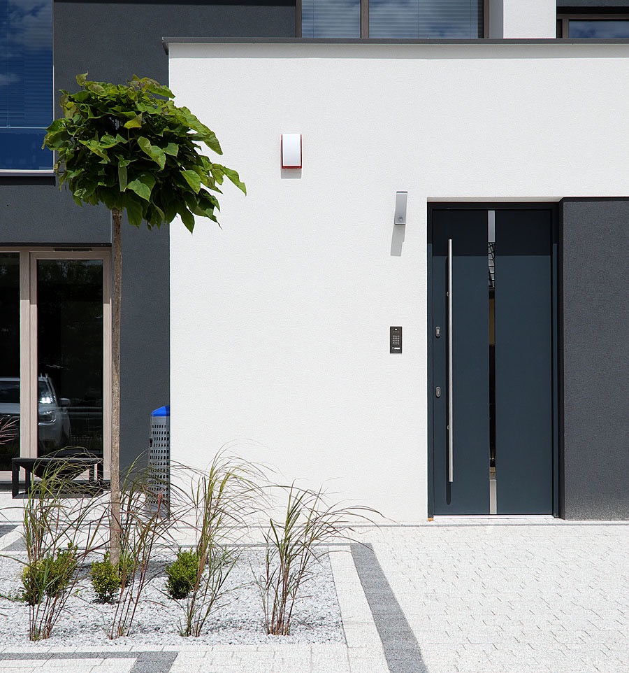 Nowoczesne wejście do domu z grafitowymi drzwiami zewnętrznymi, kodowanym zamkiem, oświetleniem elewacyjnym i ozdobnymi trawami na tle białej elewacji.