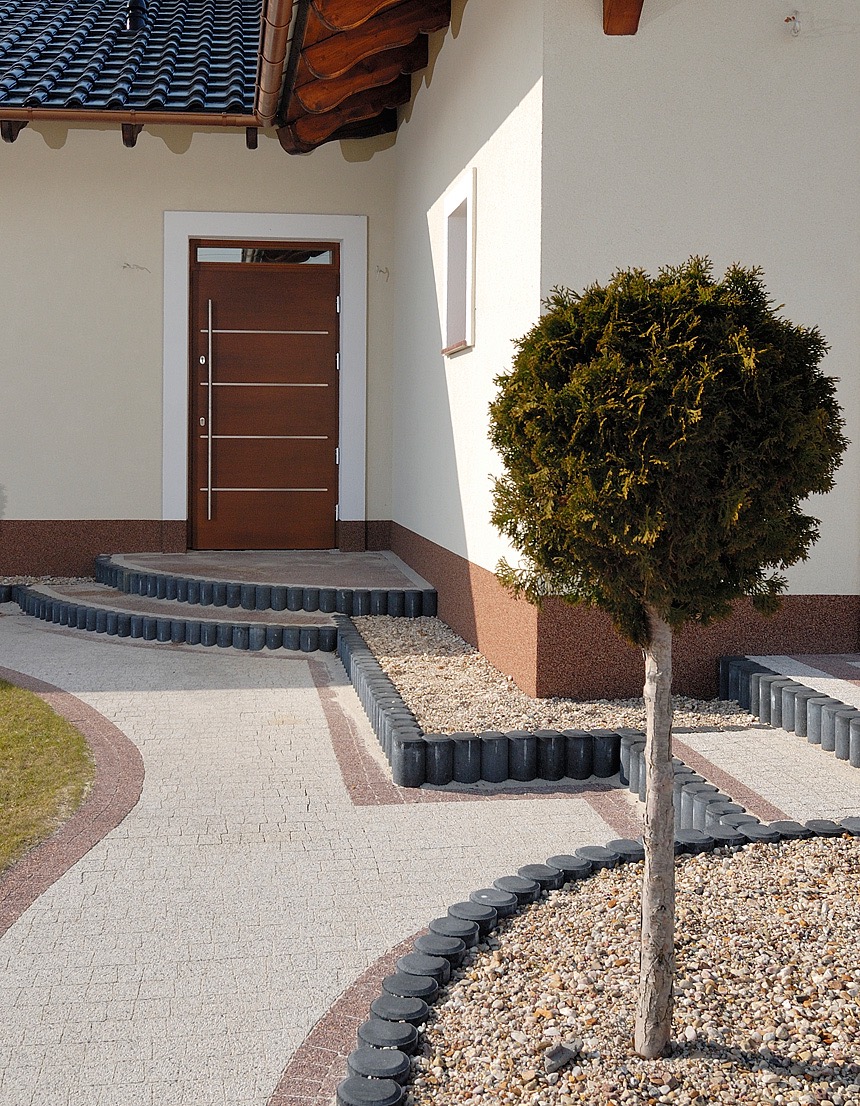 Brązowe, nowoczesne wejście do domu w Szydłowicach z geometrycznymi zdobieniami, widoczne schody z kostki brukowej i ozdobny krzew.