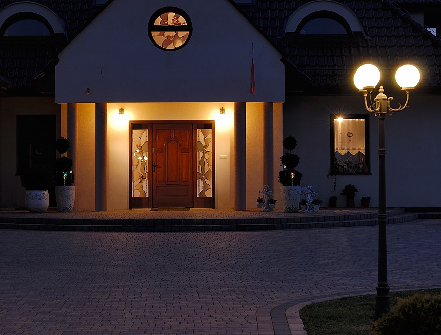 Wejście do domu jednorodzinnego oświetlone wieczornym światłem, z drewnianymi drzwiami z dekoracyjnymi szybami i lampą uliczną.