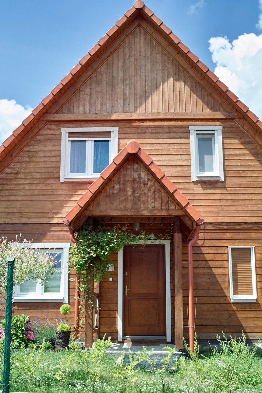 Drewniany dom z brązowymi drzwiami wejściowymi, porośniętymi zielonym bluszczem, widok z przodu, słoneczny dzień.