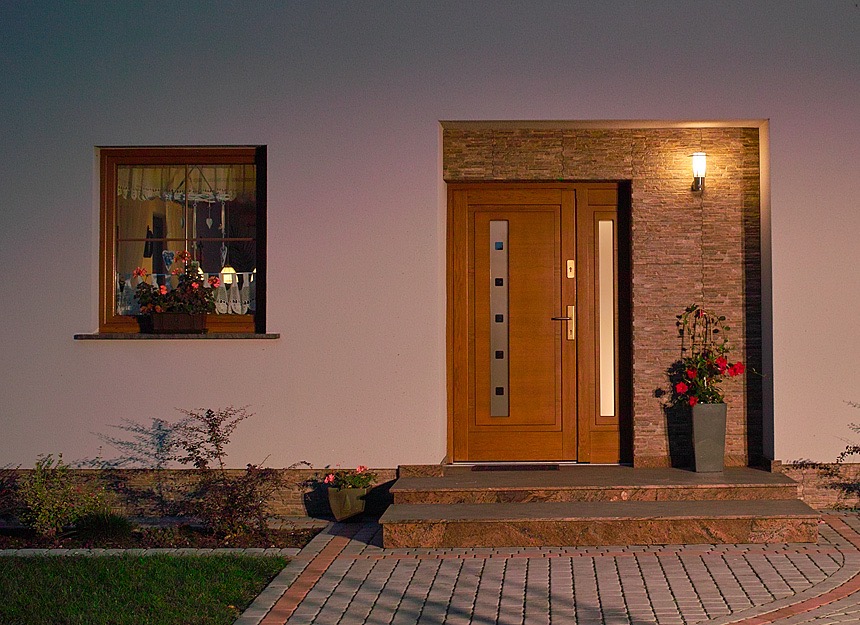 Zewnętrzne drzwi wejściowe z drewnianą ramą i przeszkleniami, oświetlone lampą ścienną, z kwiatami w donicach na stopniach i w oknie, widoczne z zewnątrz domu.