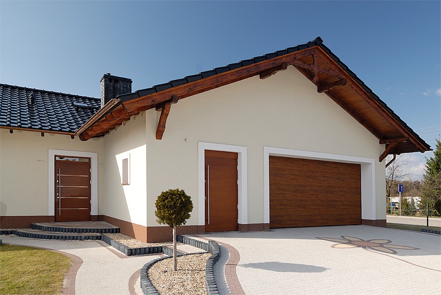 Elewacja domu z drewnianą bramą garażową i nowoczesnymi drzwiami wejściowymi z poziomymi, metalowymi akcentami, na tle jasnego nieba i kostki brukowej z motywem kwiatowym.